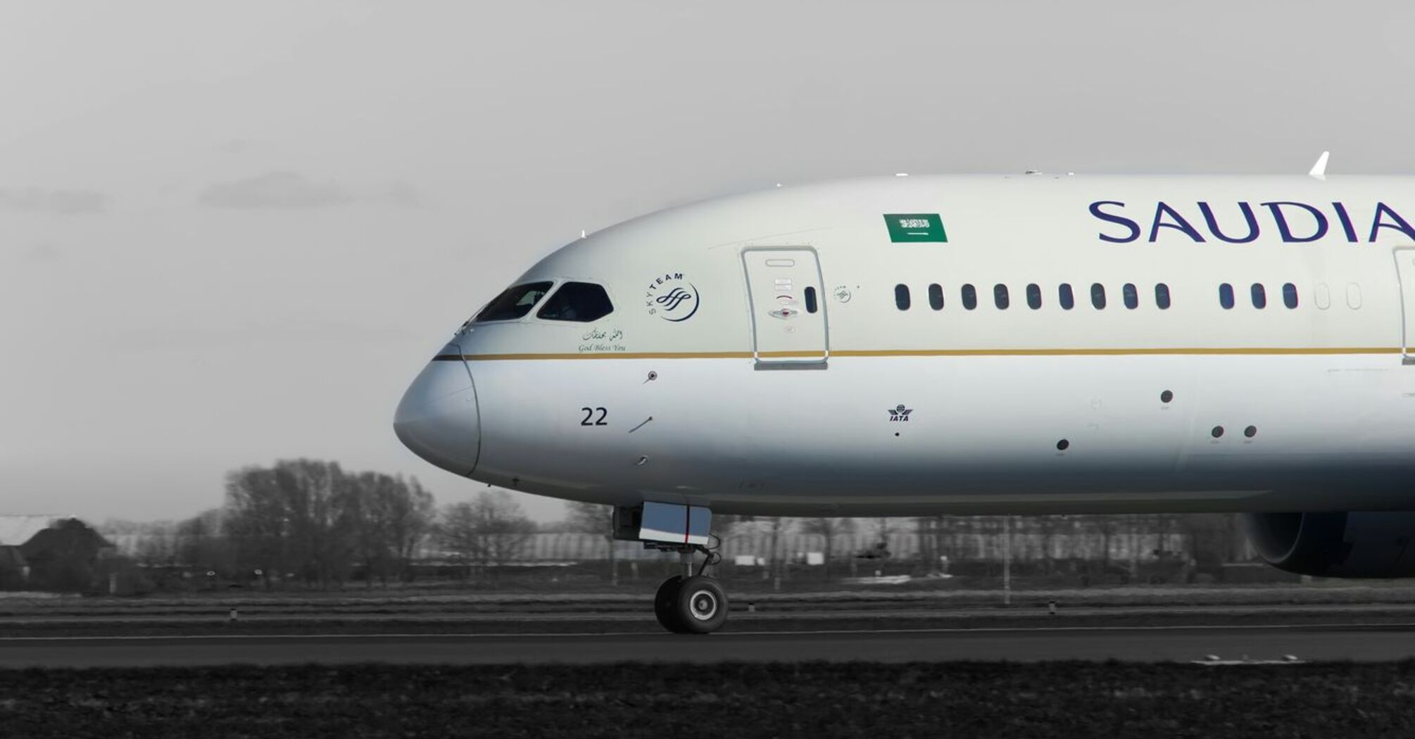 A Saudia Boeing 787 aircraft on the runway, side view, with a grayscale background highlighting the plane's livery 