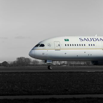 A Saudia Boeing 787 aircraft on the runway, side view, with a grayscale background highlighting the plane's livery