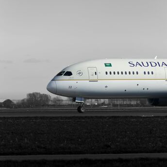A Saudia Boeing 787 aircraft on the runway, side view, with a grayscale background highlighting the plane's livery 