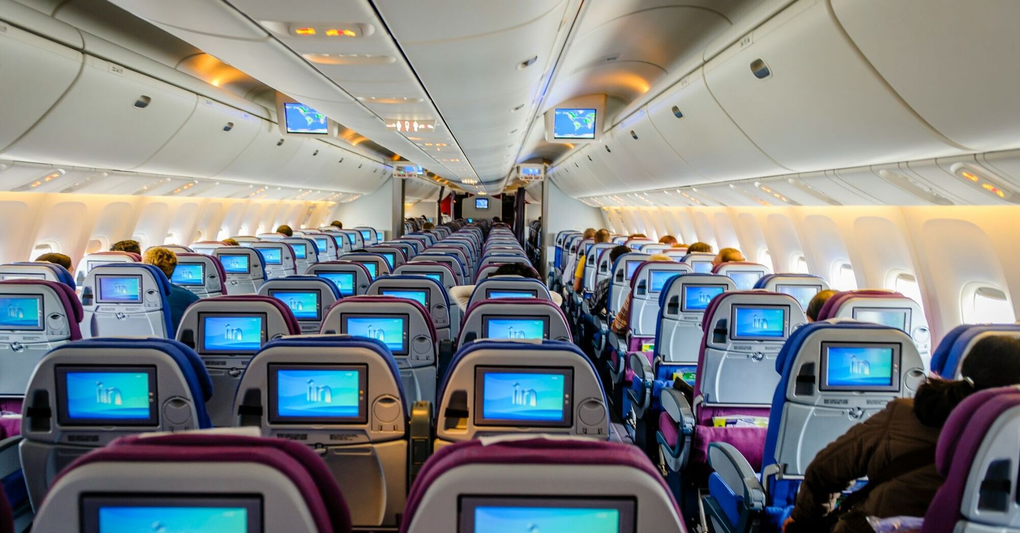 Interior of an airplane showing rows of seats with personal entertainment screens illuminated, cabin overhead lighting, and passengers seated 
