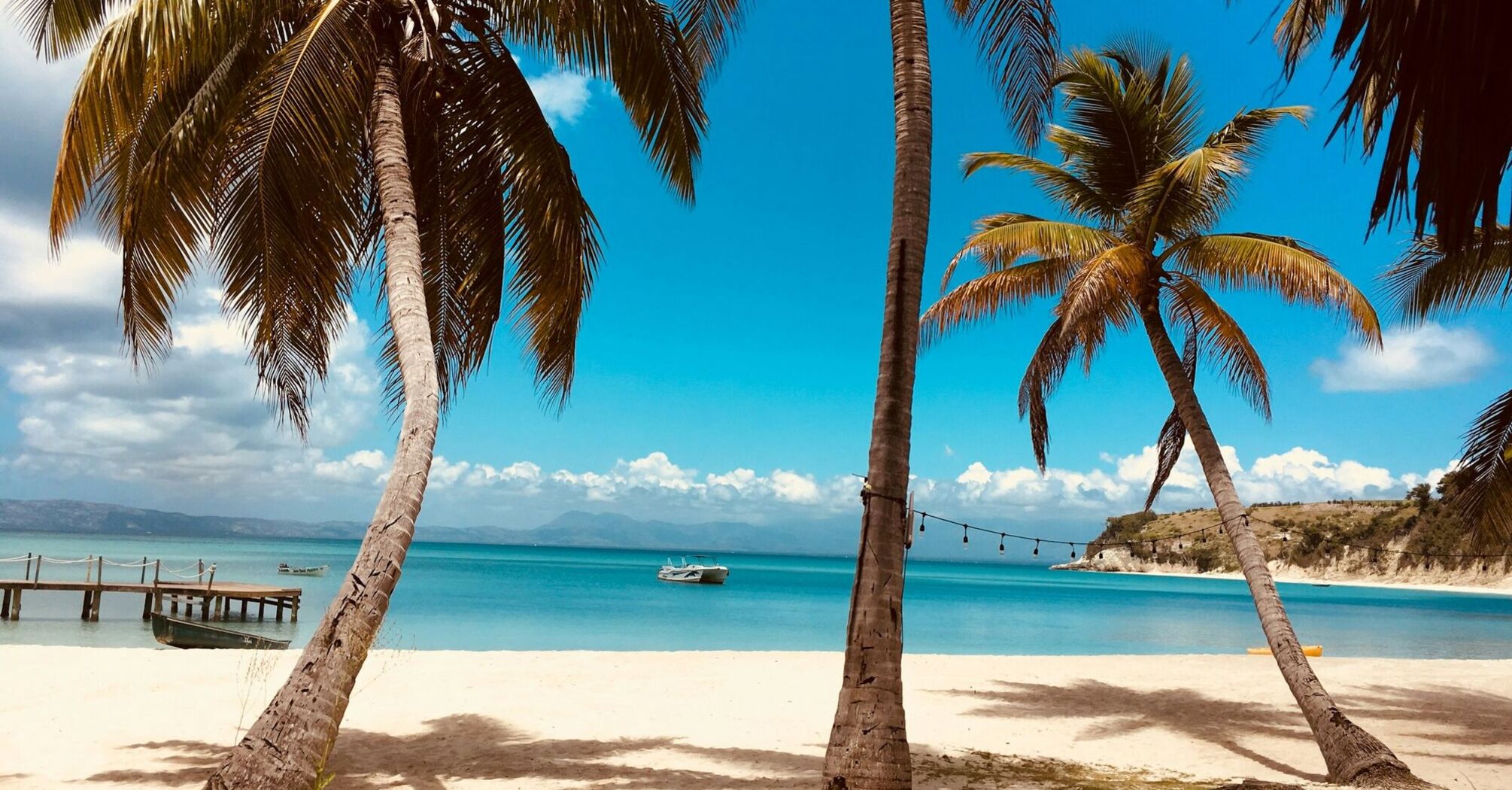 Tropical beach view with clear blue sky, palm trees on the sandy shore, a calm sea, and a small boat near a wooden pier in the distance