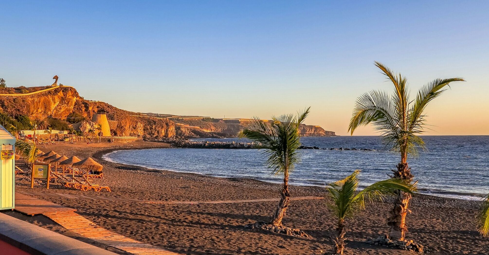View of a tranquil beach in Tenerife with palm trees in the foreground and cliffs in the distance during sunset