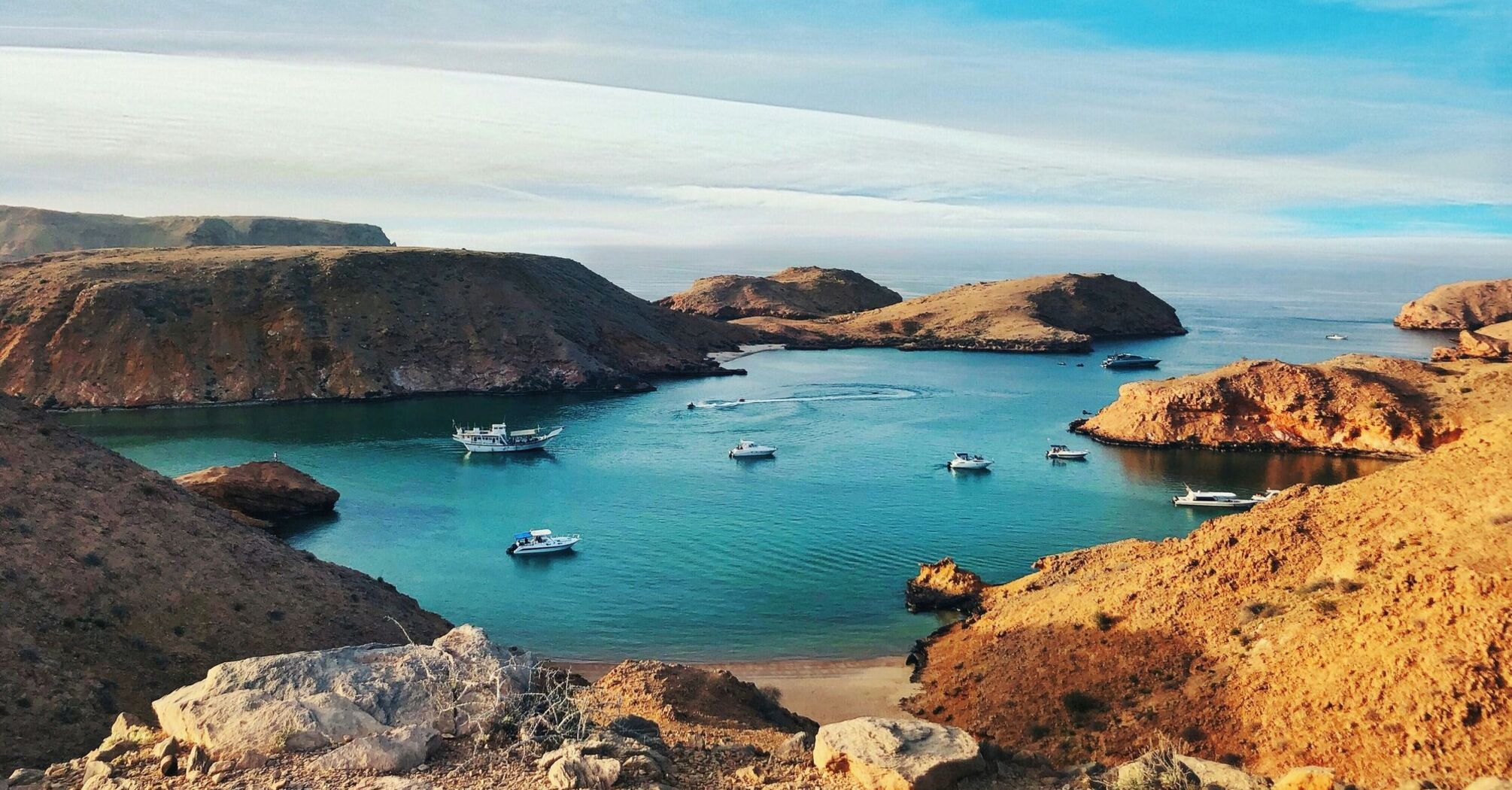 Picturesque view of a serene bay with boats on the clear waters of the Gulf of Oman, surrounded by rugged hills under a blue sky