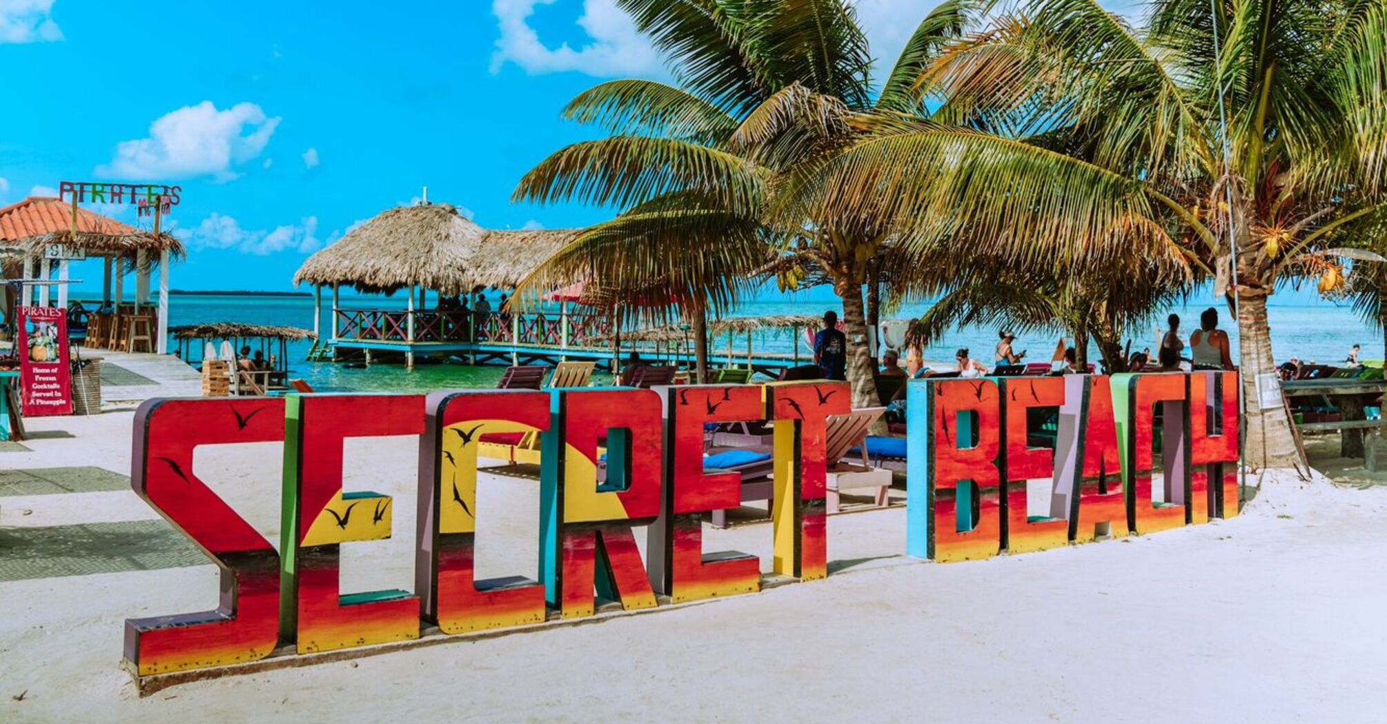 Colorful 'SECRET BEACH' sign with palm trees and beach huts in the background under a clear blue sky at Secret Beach, San Pedro, Belize
