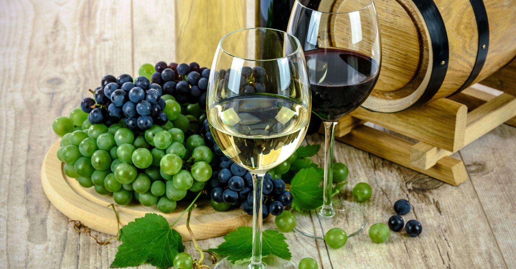 A glass of white wine and a glass of red wine with bunches of green and blue grapes, leaves, a wine bottle, and a wooden barrel on a wooden surface