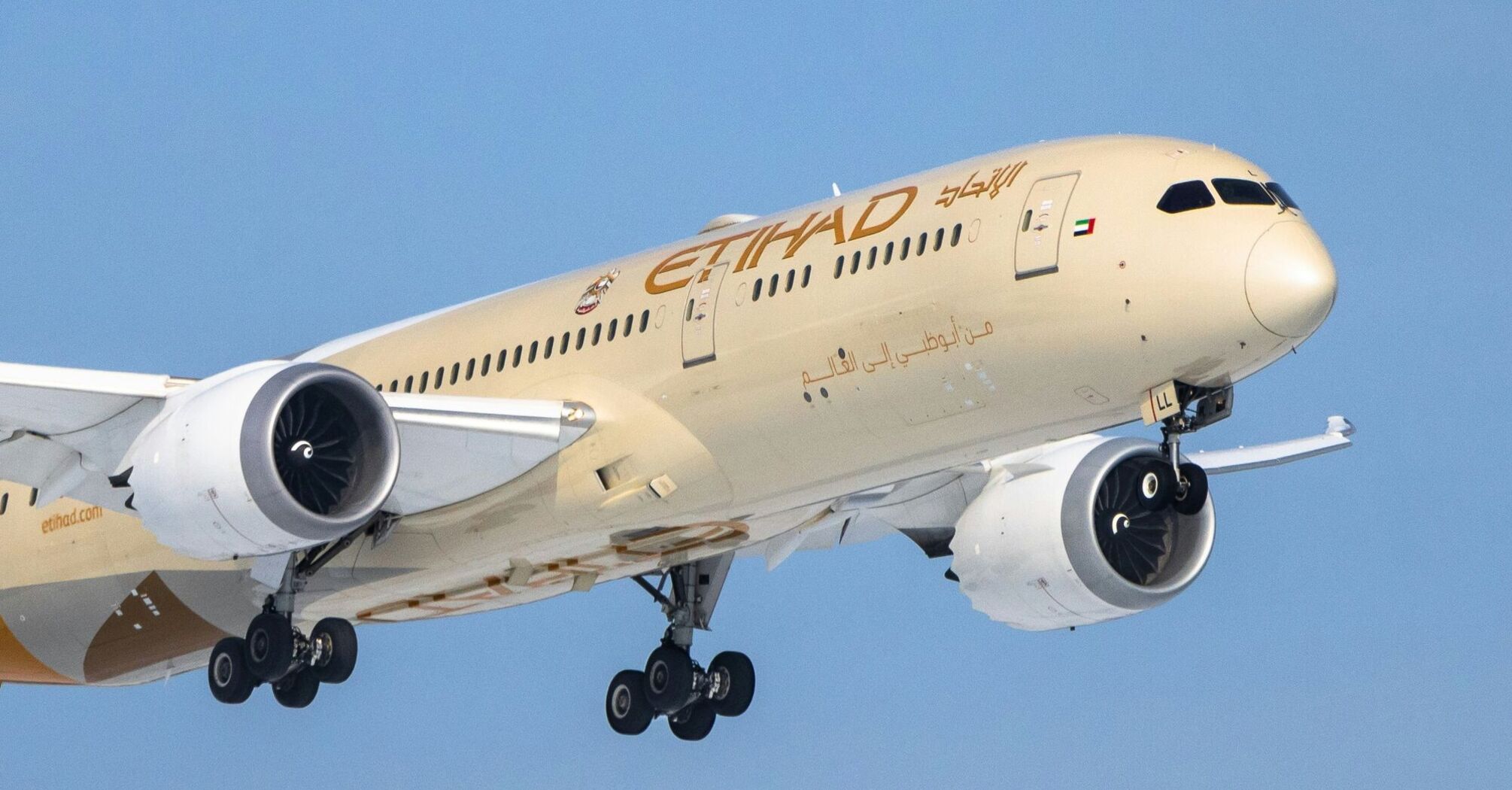 An Etihad Airways aircraft in mid-flight, with landing gear extended and the golden livery featuring the airline's name both in English and Arabic script