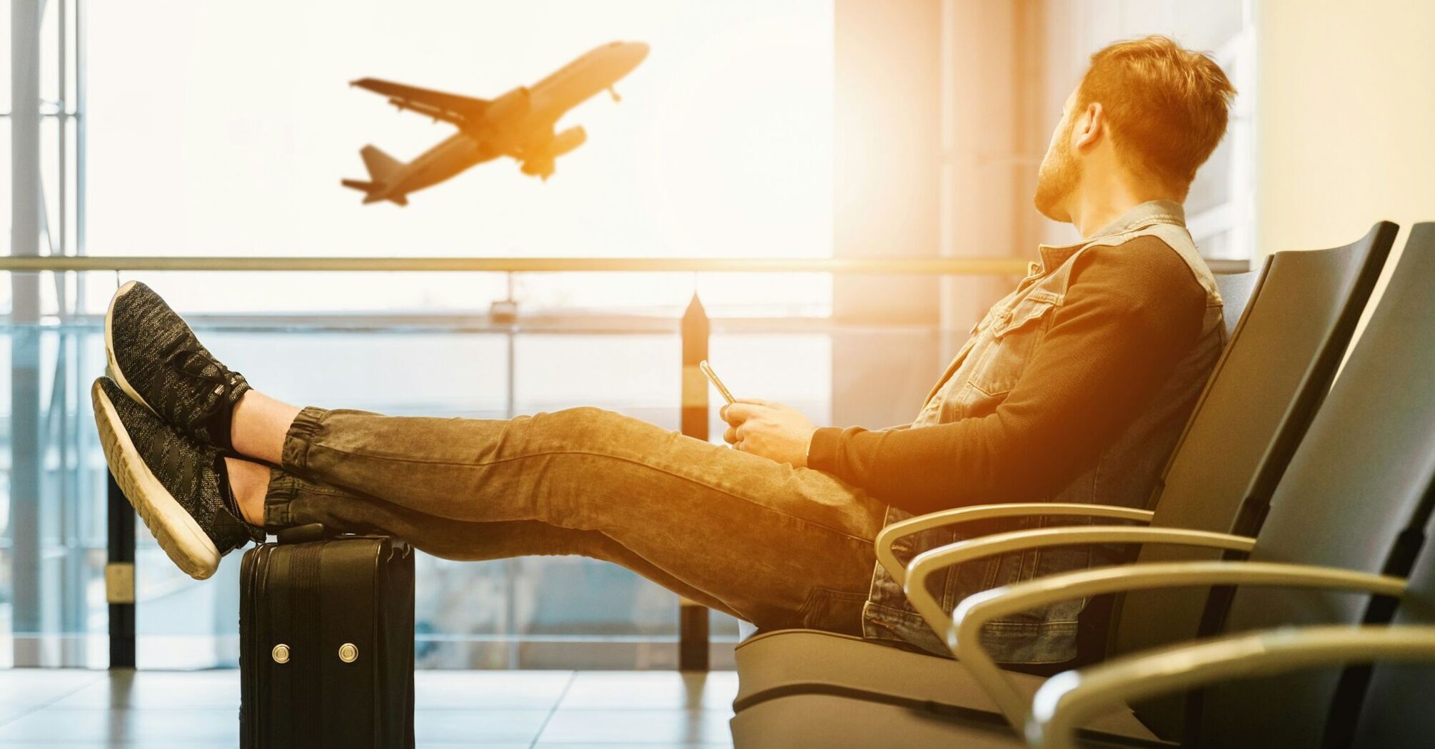 A traveler sitting relaxed in an airport lounge with feet up on a suitcase, looking at a smartphone, with an airplane taking off in the background through the window