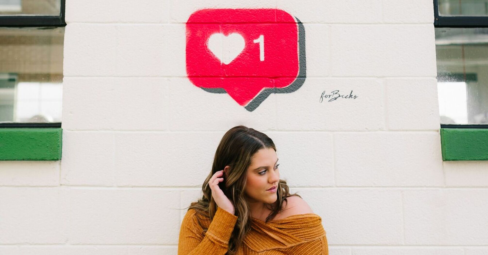 A woman in a mustard yellow sweater standing against a white wall with a painted red social media 'like' notification icon