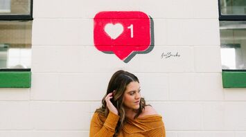 A woman in a mustard yellow sweater standing against a white wall with a painted red social media 'like' notification icon
