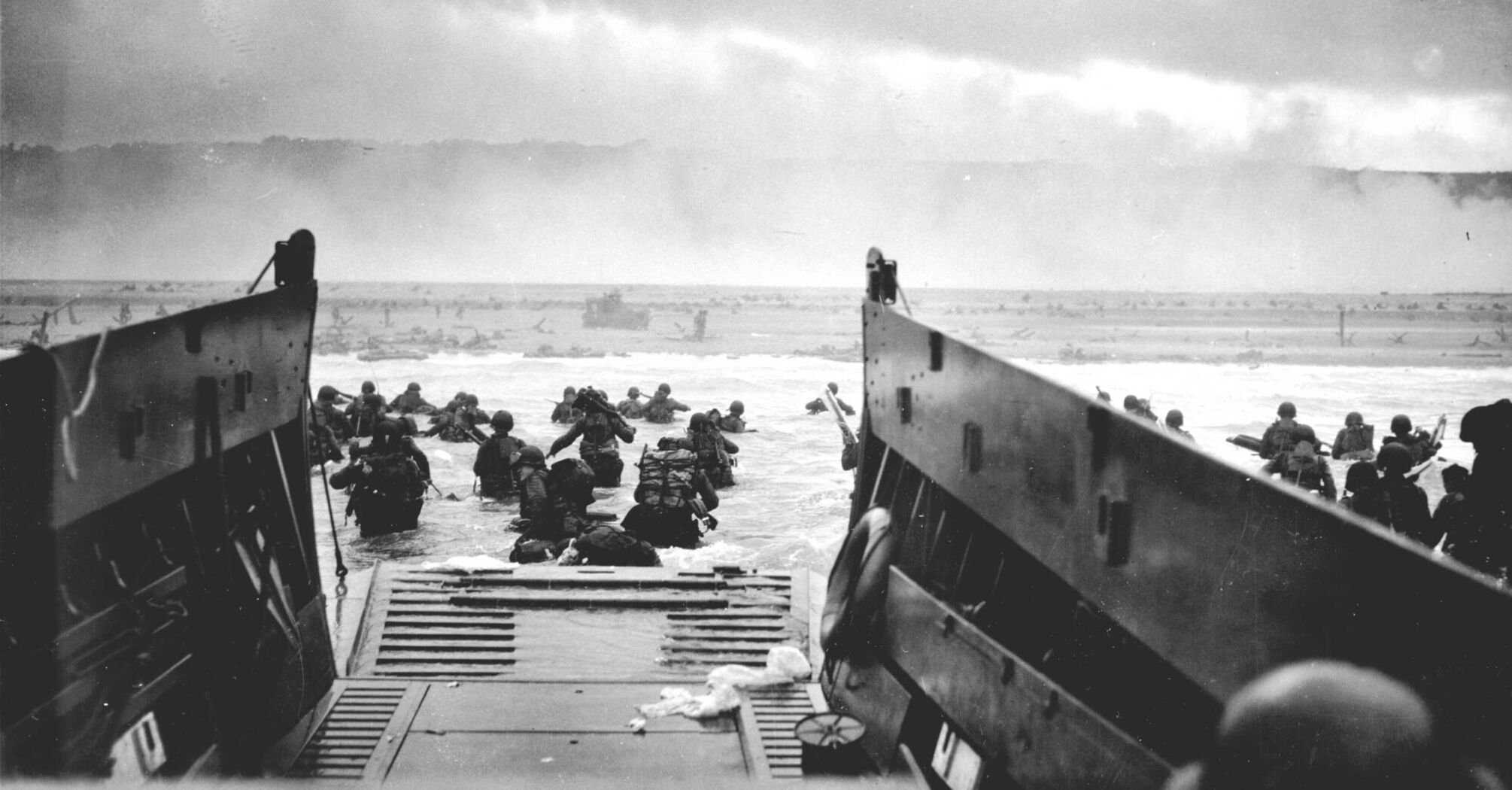 Black and white photograph of the landing of Allied troops on the beach during D-Day, with soldiers disembarking from landing craft into the sea under fire 