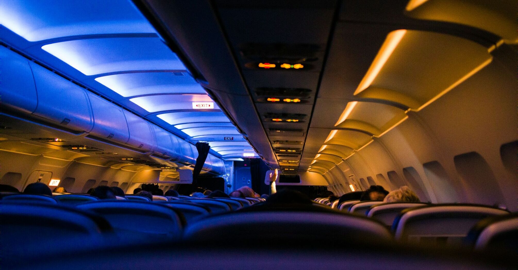 The interior of an airplane cabin at night with ambient blue overhead lighting and rows of unoccupied seats leading towards the exit sign 