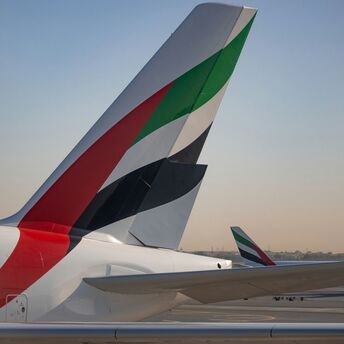 Тails of Emirates aircraft with iconic red, black, white, and green design, on the tarmac with a clear sky in the background