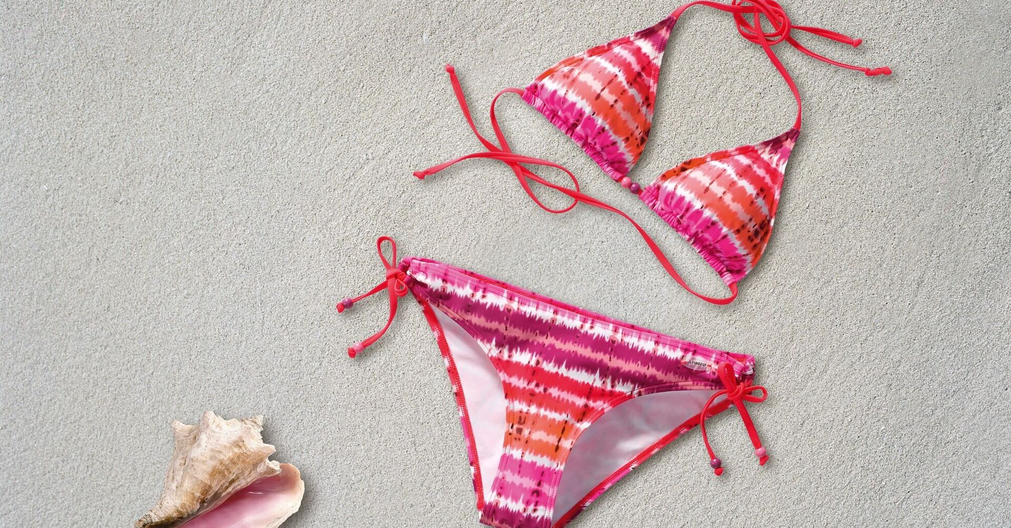 A pink and orange tie-dye bikini with string ties, laid out on a sandy surface alongside a conch shell and a seashell