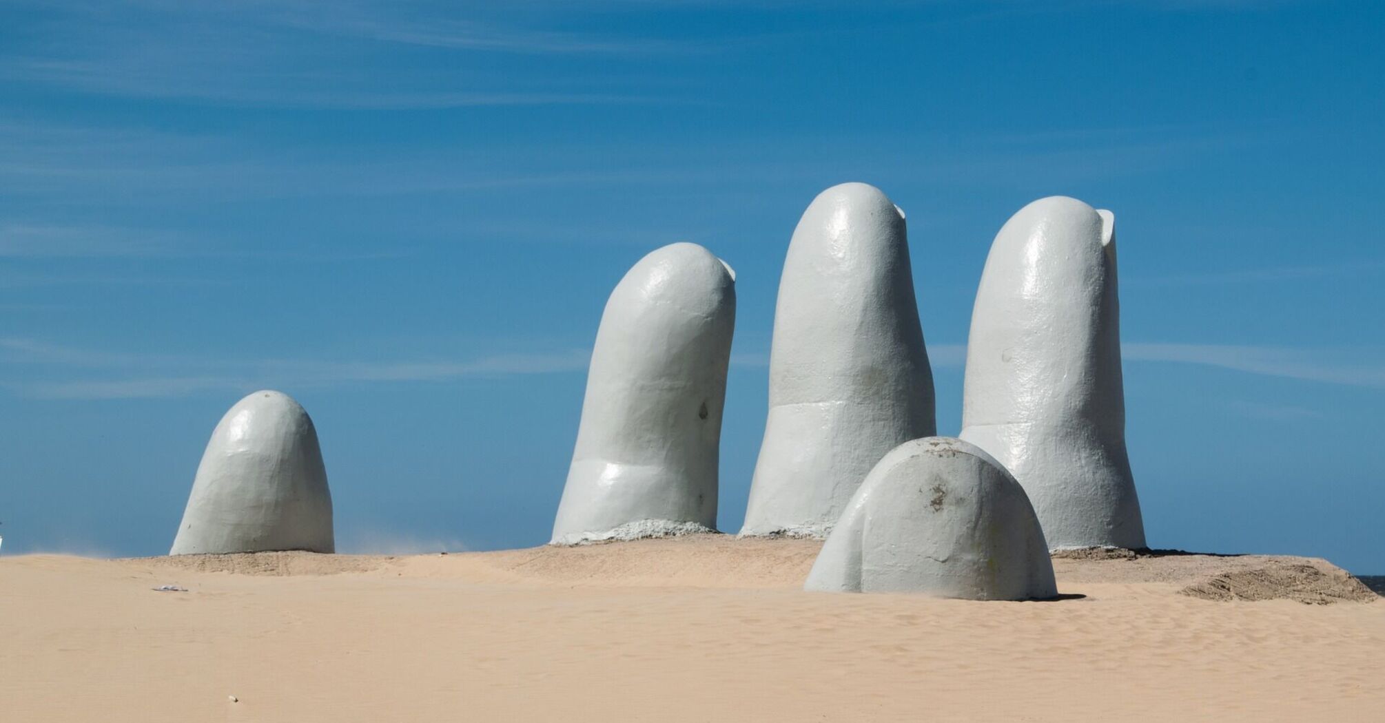 A large sculpture of a hand partially buried in sand on a beach under a clear blue sky 