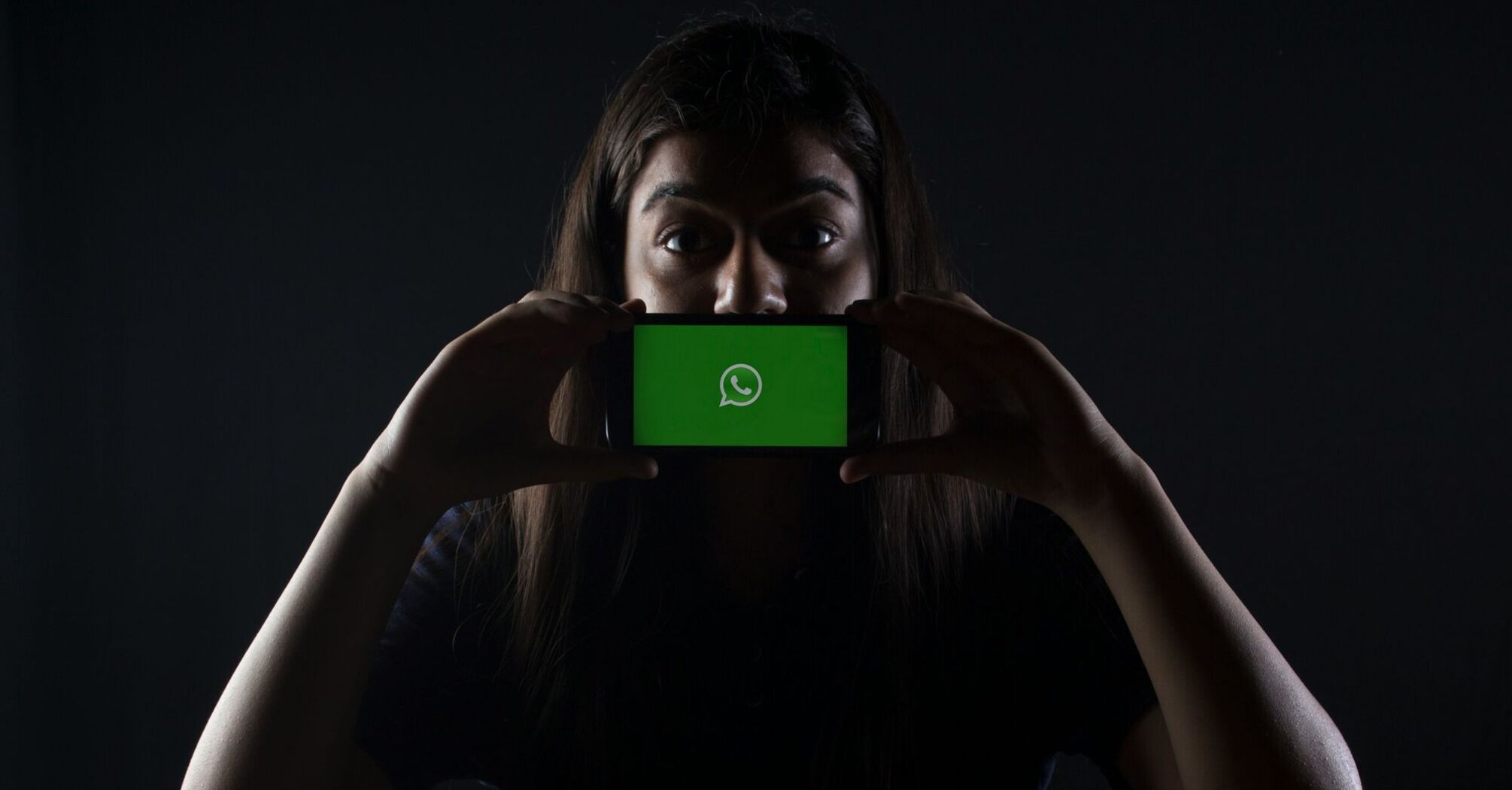 A person holding a smartphone horizontally in front of their face, displaying the WhatsApp logo on the screen, with a dark background