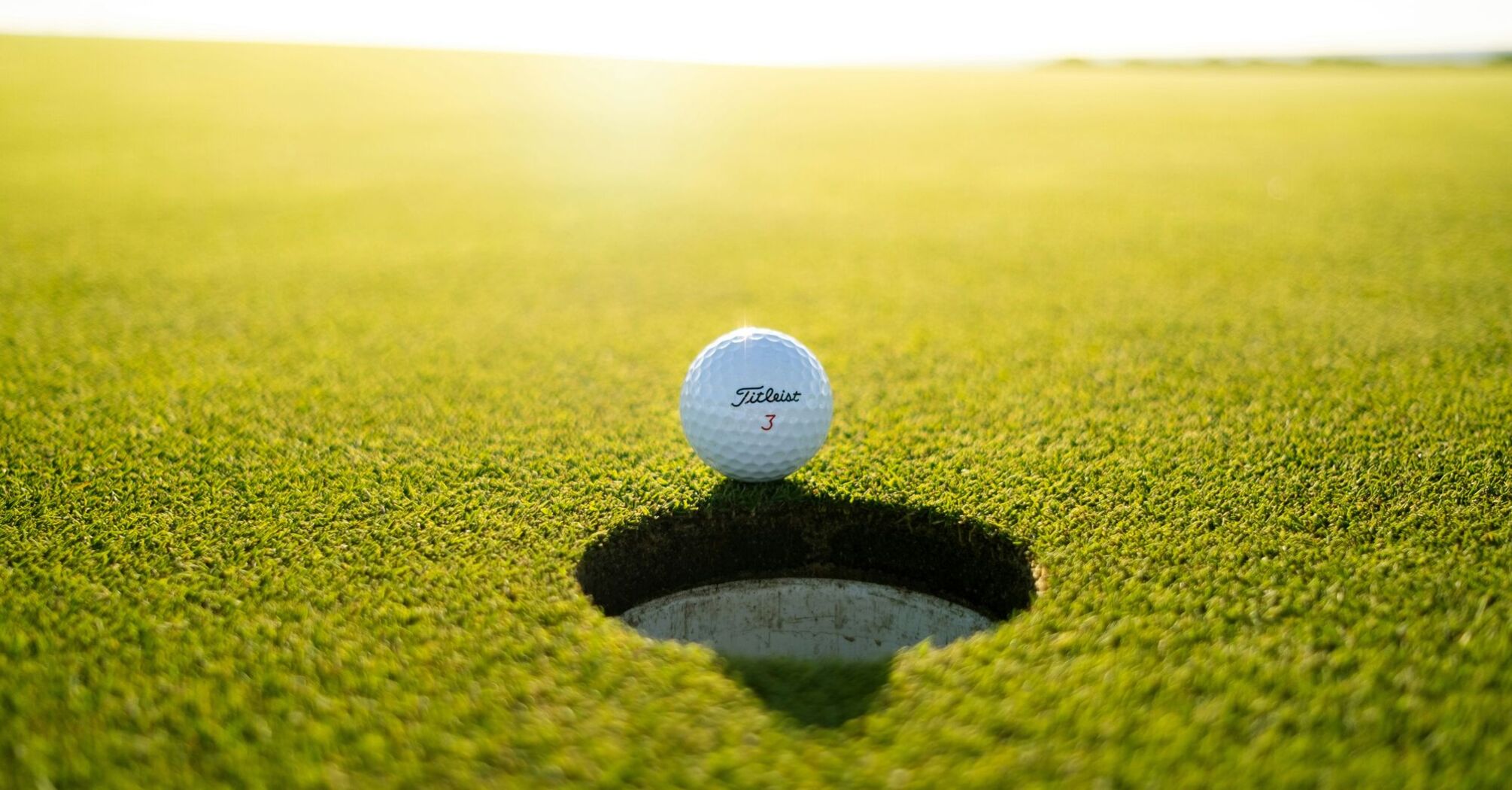 A golf ball on the edge of the hole on a vibrant green golf course, with sunlight casting a gentle glow across the horizon