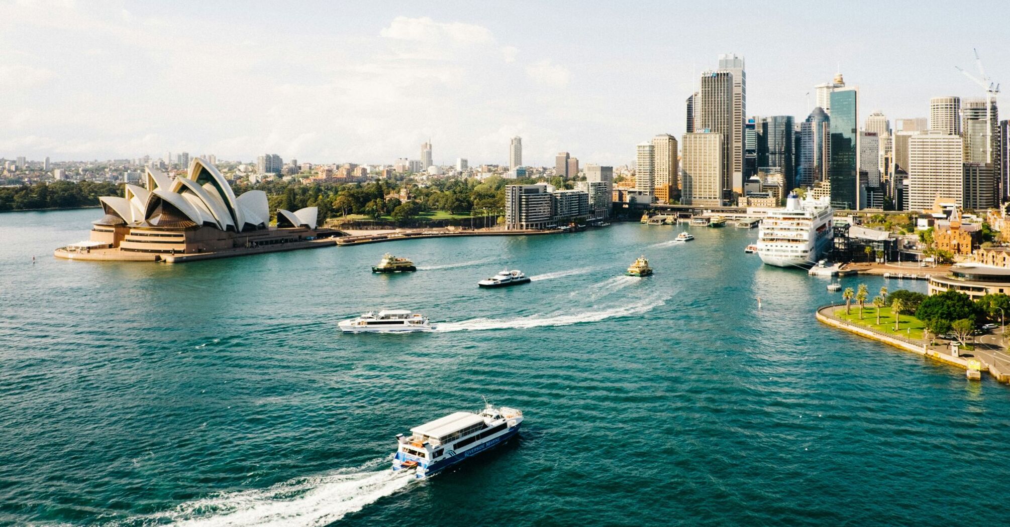 A panoramic view of Sydney Harbour, featuring the iconic Sydney Opera House, surrounded by the city skyline and various boats on the water