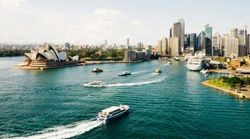 A panoramic view of Sydney Harbour, featuring the iconic Sydney Opera House, surrounded by the city skyline and various boats on the water
