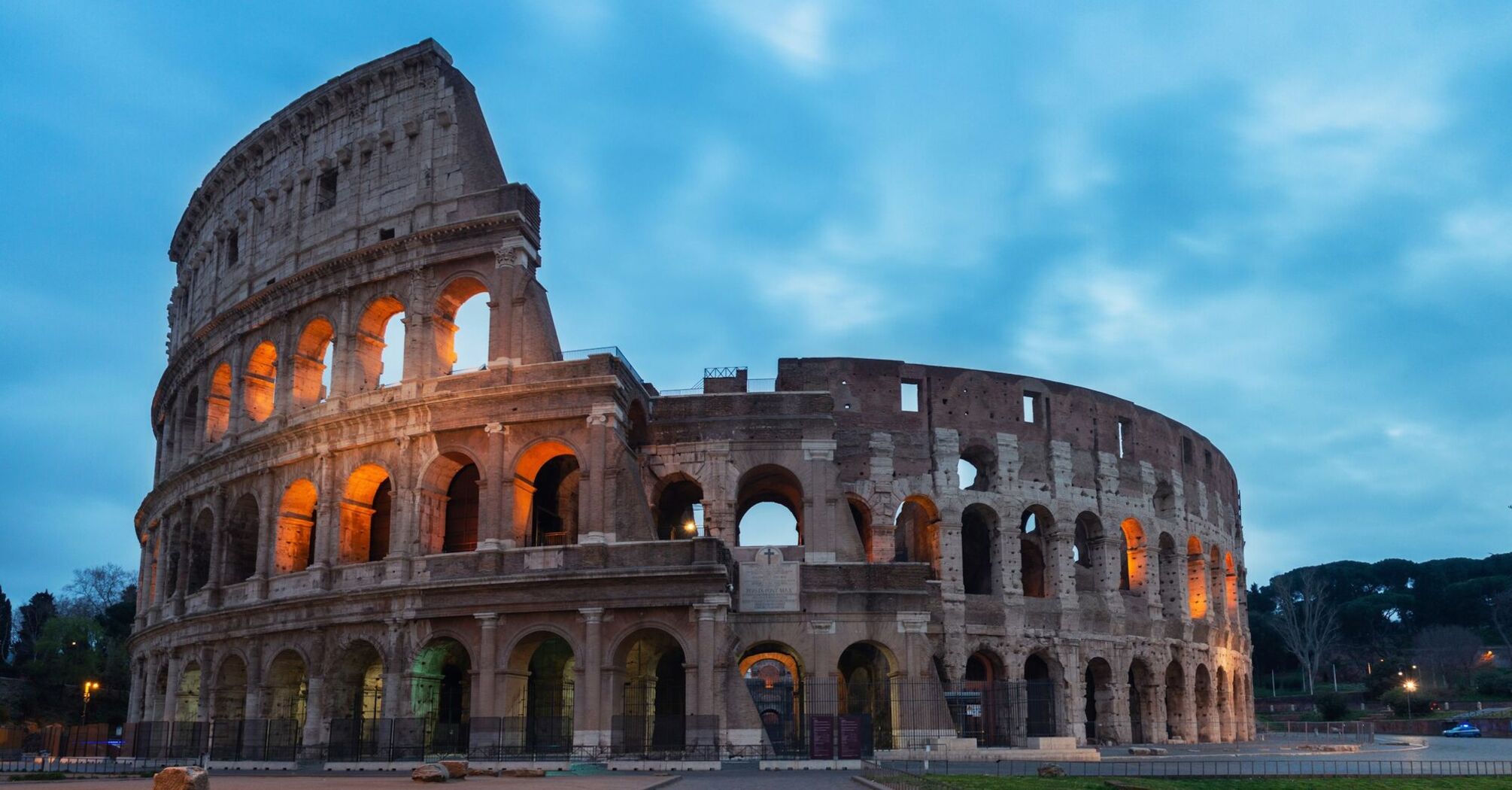 The Colosseum in Rome illuminated at dusk with a cloudy blue sky in the background