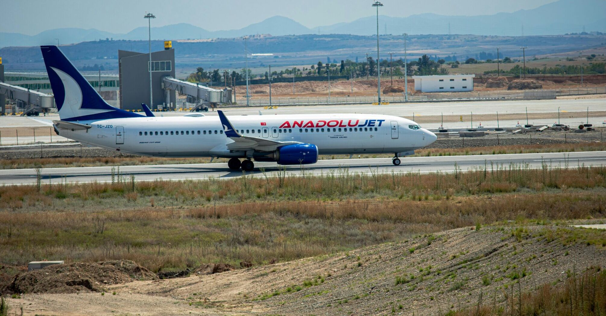 An AnadoluJet aircraft on the tarmac at an airport with airport infrastructure in the background and an open field in the foreground
