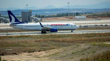 An AnadoluJet aircraft on the tarmac at an airport with airport infrastructure in the background and an open field in the foreground