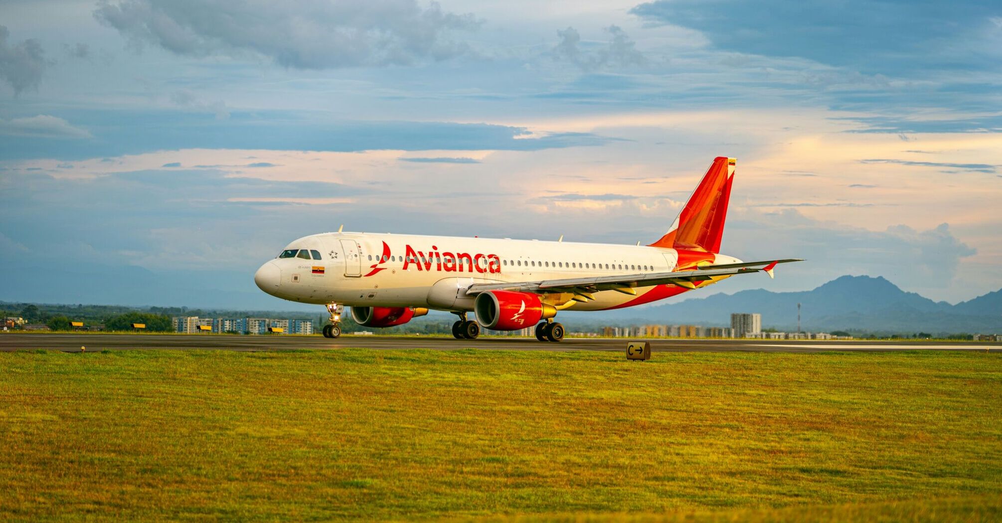 An Avianca airplane is taxiing on the runway with a backdrop of a scenic, cloud-streaked sky and distant mountains