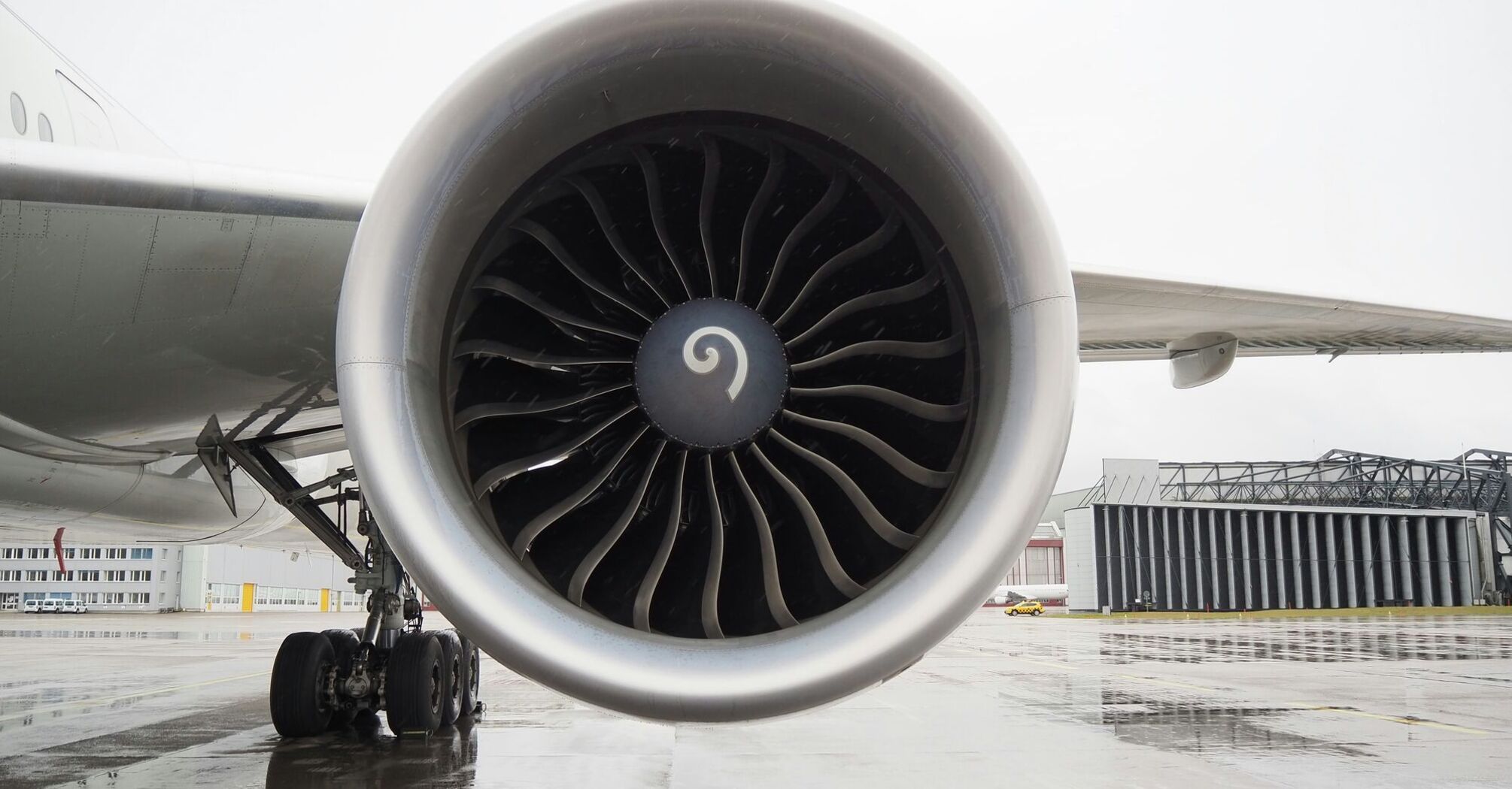 Close-up view of a jet engine on an aircraft wing, parked at an airport on a wet tarmac with airport structures in the background