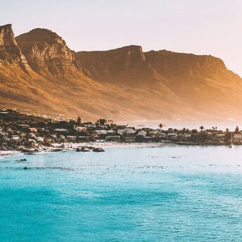 The warm glow of sunset over Cape Town, with the Twelve Apostles Mountain Range overlooking the calm blue waters and sandy beach