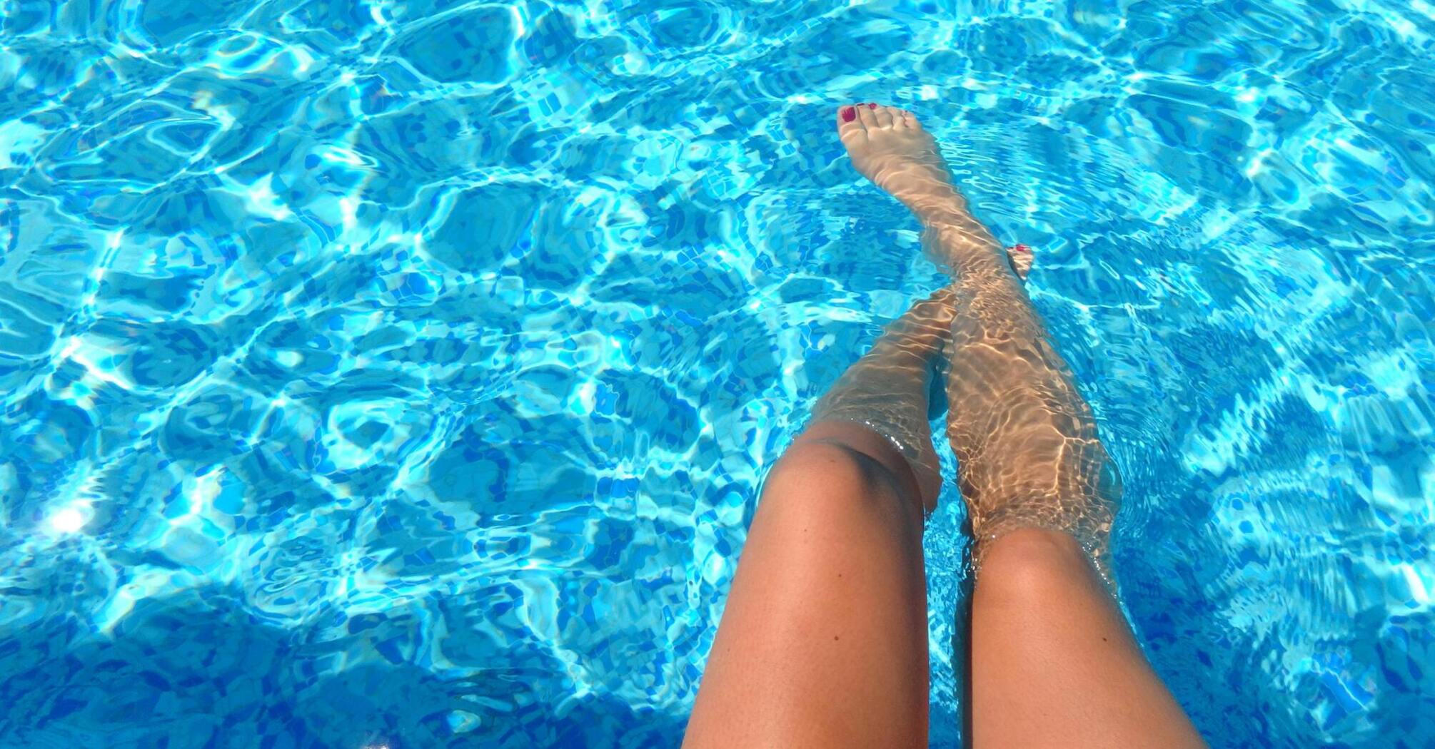 Person's legs dangling in clear blue pool water, with sunlight reflecting off the ripples 
