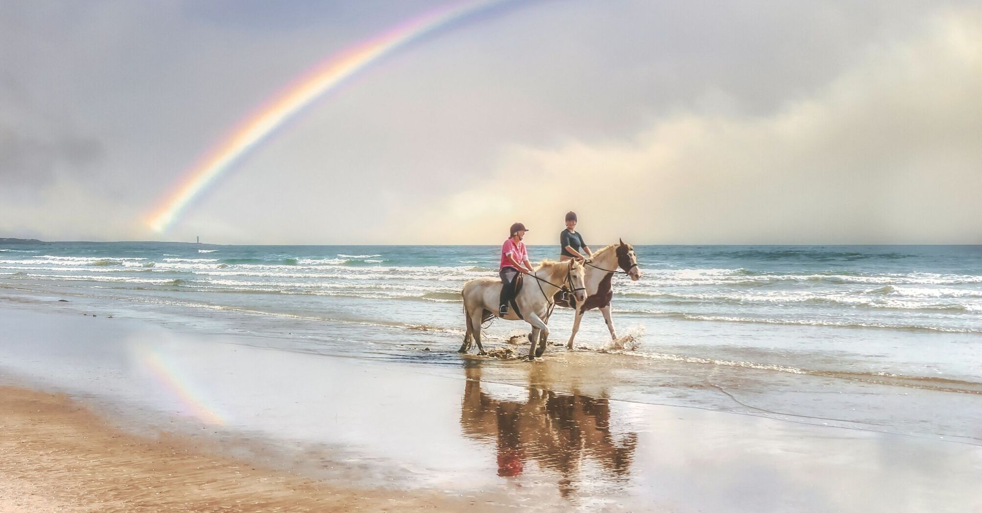 Two people riding horses on a beach with a vibrant rainbow in the background 
