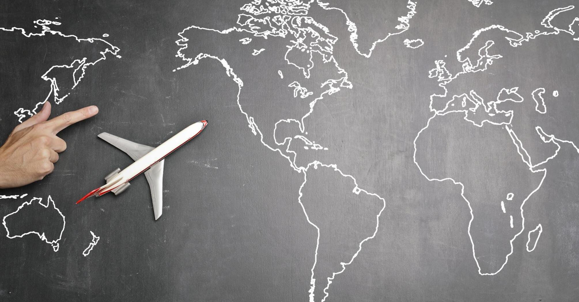 A person's hand pointing at a chalk-drawn world map on a blackboard, with a model airplane placed on the board
