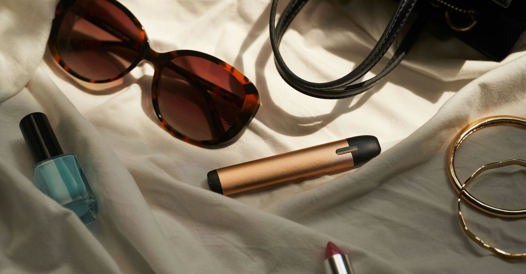 A flat lay of various personal items, including a pair of oversized sunglasses, a black handbag, a bottle of nail polish, a lipstick, and golden bangles