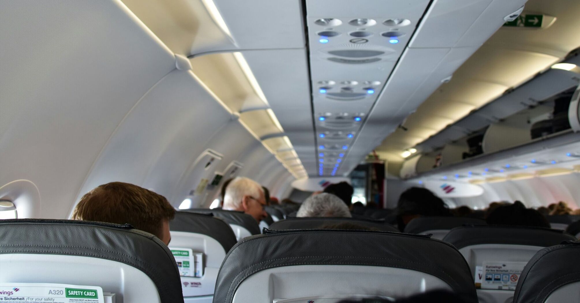 An airplane cabin interior viewed from the perspective of a passenger, showing the backs of seats, overhead compartments, and ceiling with air vents and lights