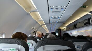 An airplane cabin interior viewed from the perspective of a passenger, showing the backs of seats, overhead compartments, and ceiling with air vents and lights