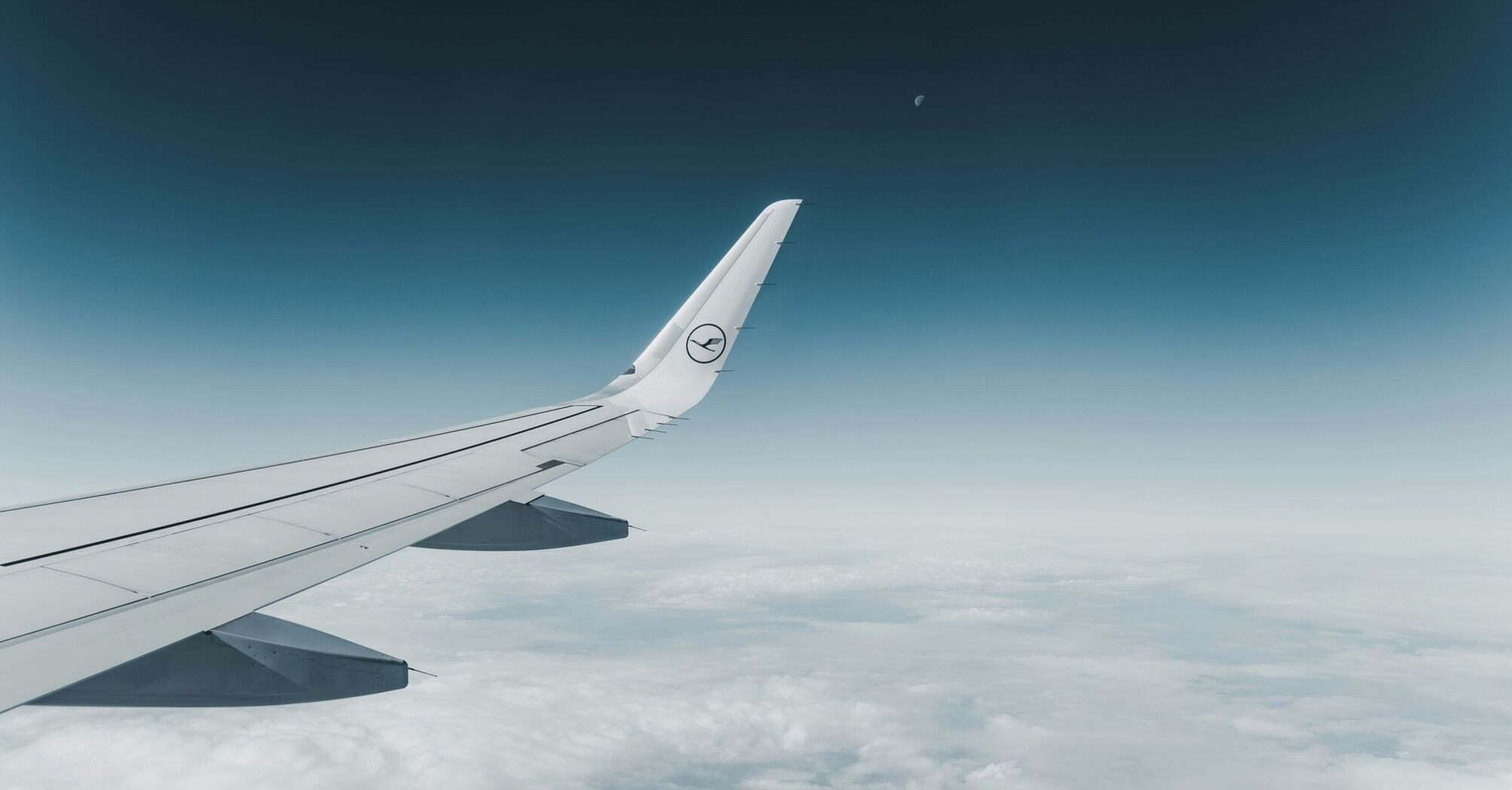 View of an airplane wing with the Lufthansa logo mid-flight against a clear sky, with a crescent moon in the distance