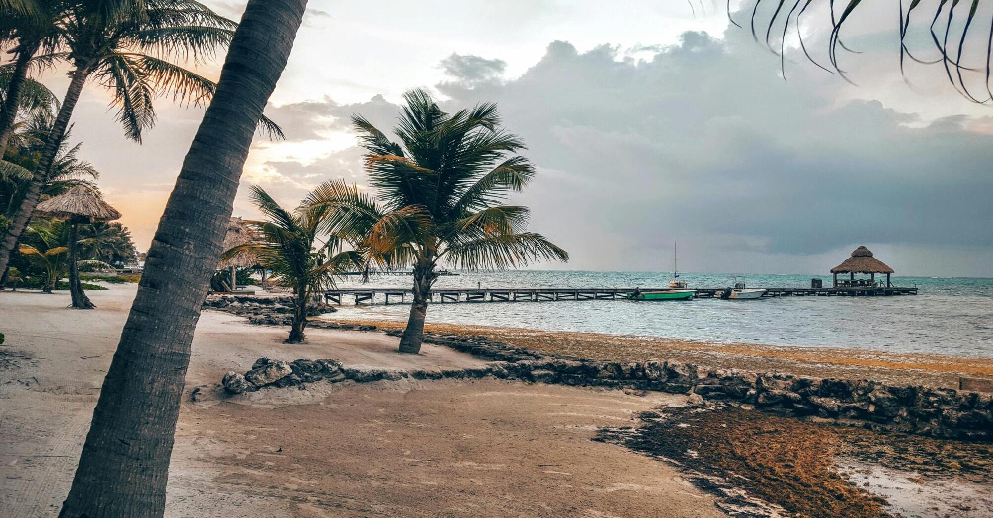 A serene view of the beach in San Pedro, Corozal District, Belize, with palm trees, a dock, and boats under a cloudy sky at dusk