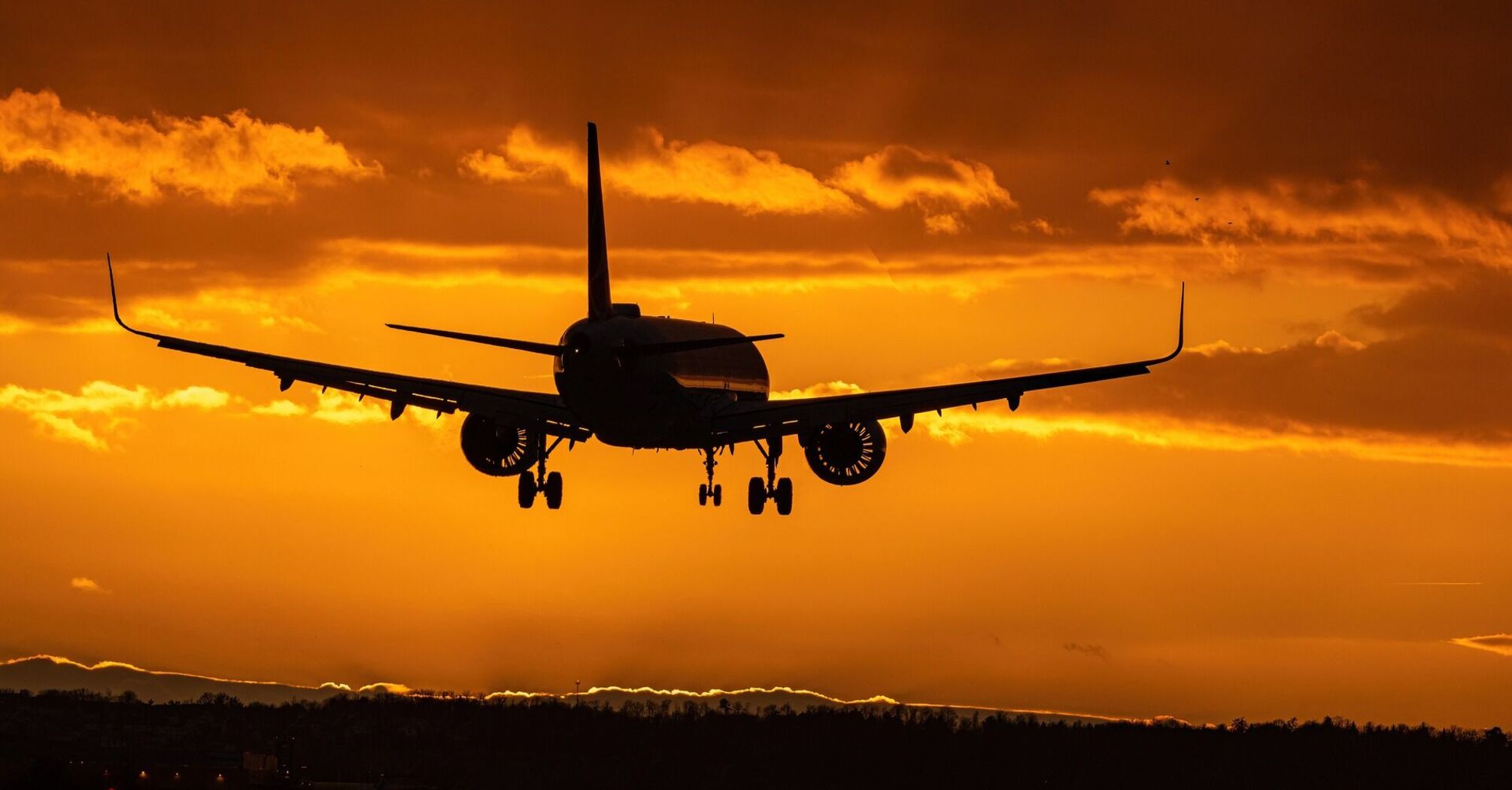 An airplane silhouetted against an orange sunset sky, preparing to land with landing gear deployed 