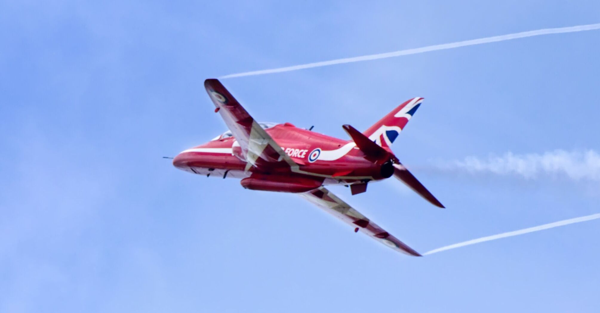 A red jet aircraft with UK insignia performing a dynamic flight maneuver against a clear blue sky with visible contrails