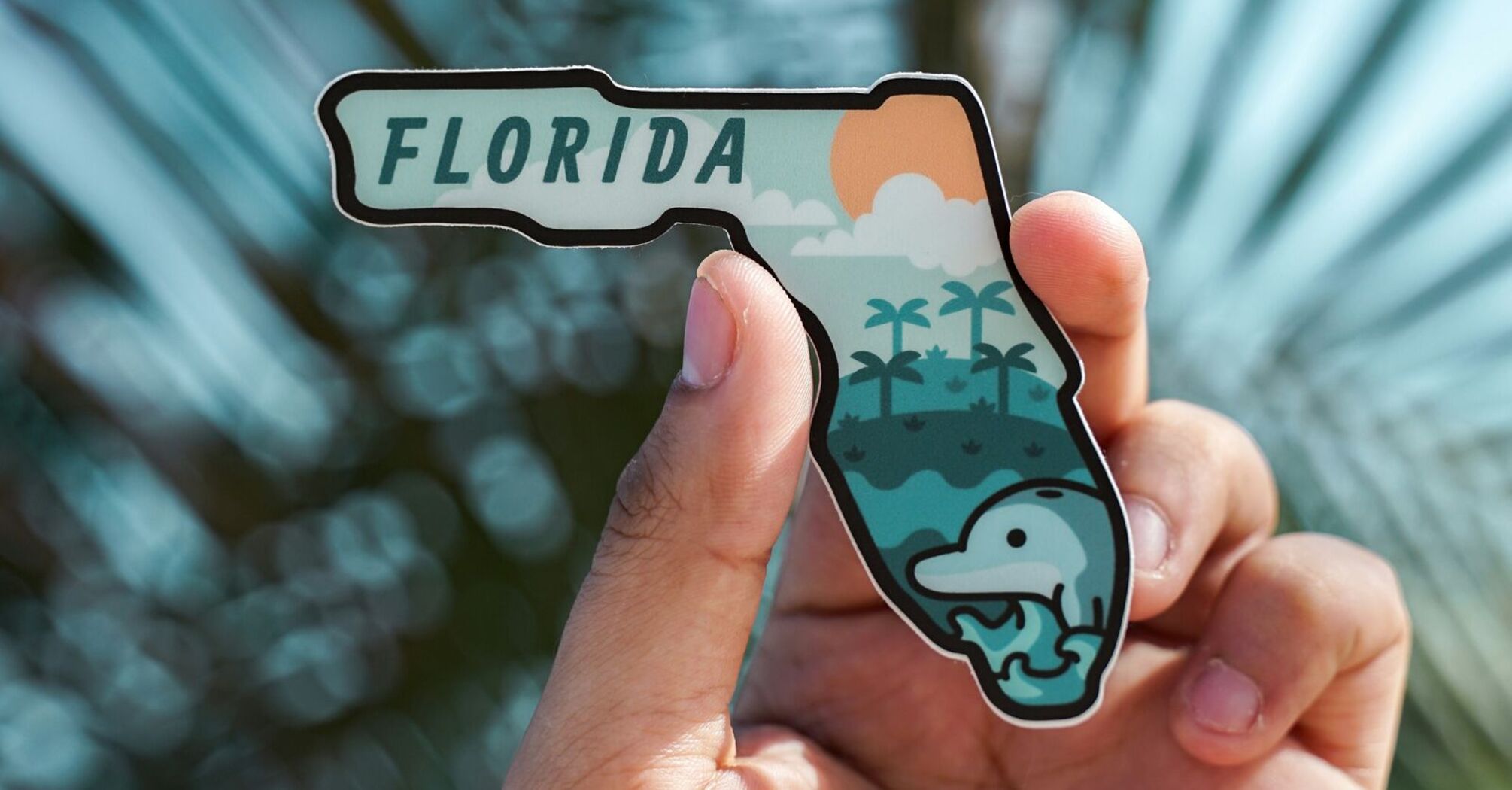 A hand holding a Florida-shaped sticker with a dolphin and palm trees design, with blurred palm fronds in the background