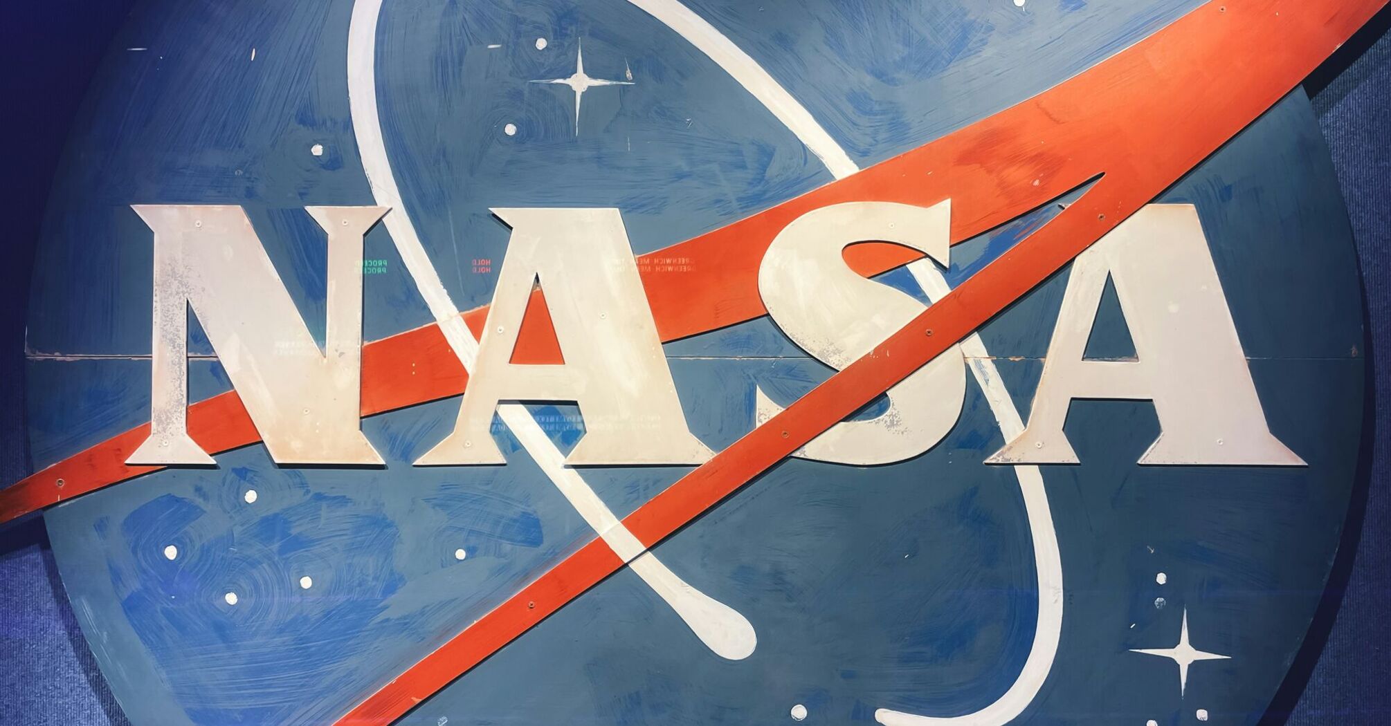 A close-up photo of a weathered NASA logo painted on a surface, featuring the iconic blue circular background with white stars, a red swoosh, and the white letters "NASA."