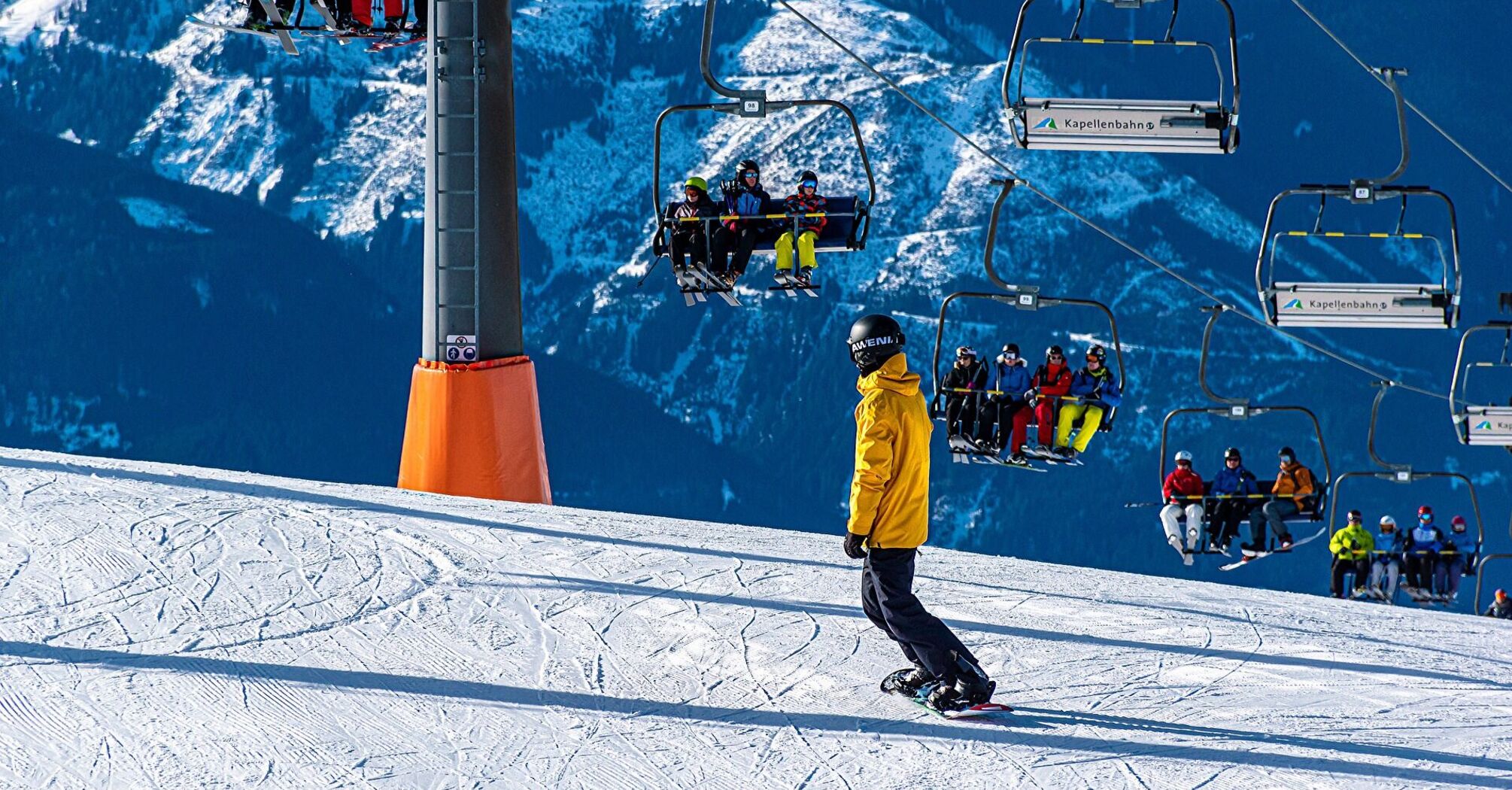 Snowboarder gliding down a snowy mountain slope with a ski lift carrying skiers in the background