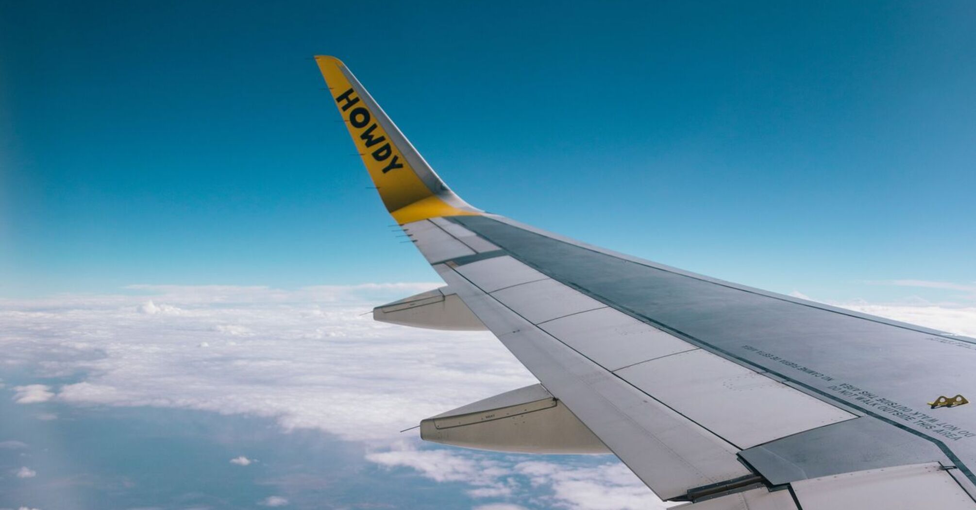 View from an airplane window showing the wing with the word 'HOWDY' written on it, flying above the clouds