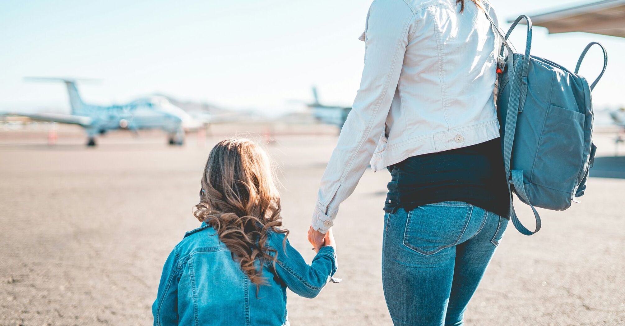 A mother and her young daughter holding hands on an airfield, with small planes in the background