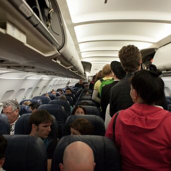 Airplane interior with passengers standing in the aisle during boarding or deplaning, showing packed overhead bins and a full cabin