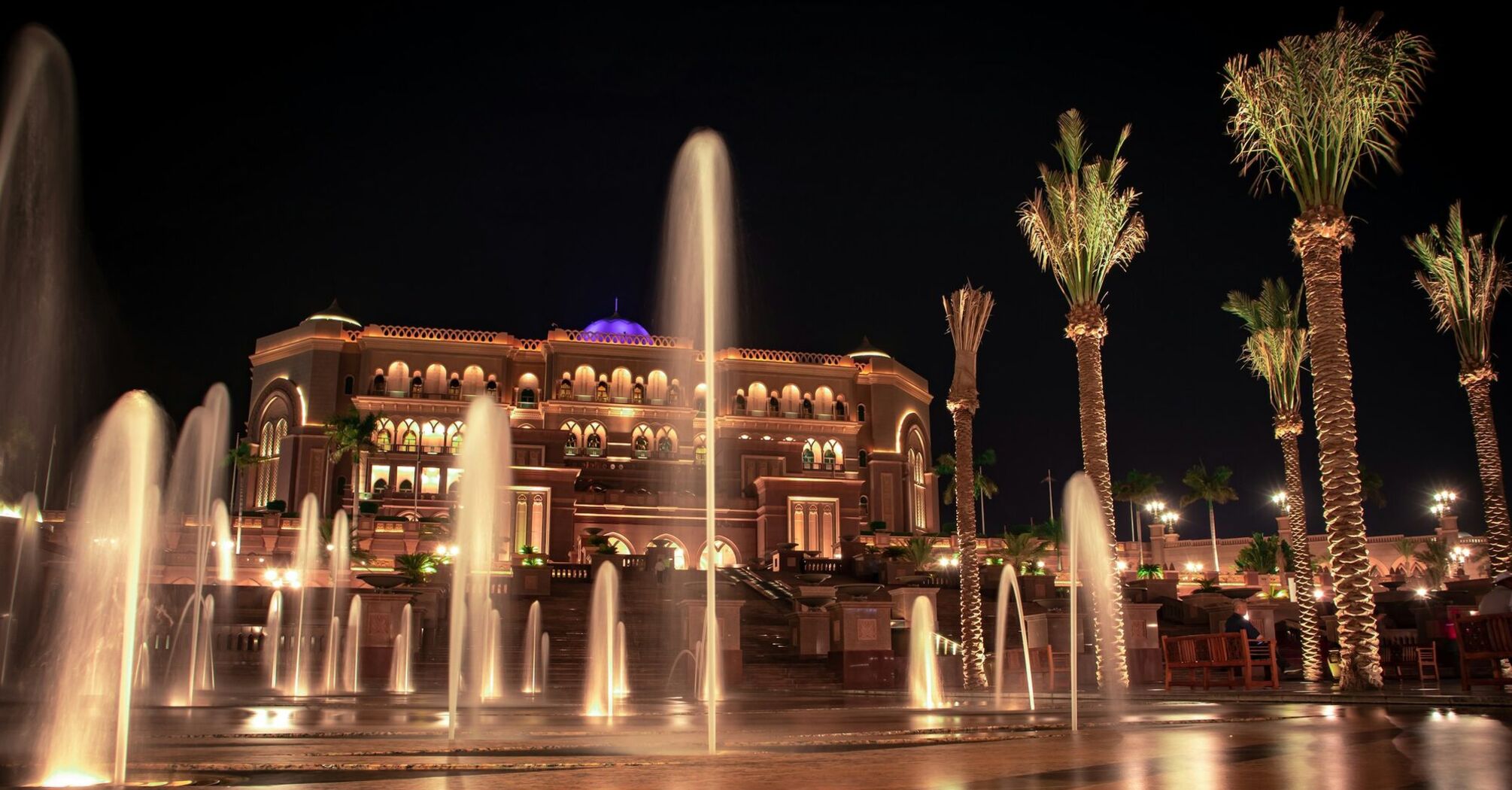 A night view of an illuminated luxurious building with arched windows and palm trees, flanked by water fountains