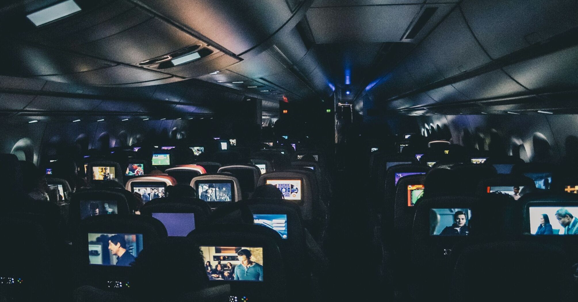 The interior of an airplane cabin with rows of seats, each with personal entertainment screens displaying various movies