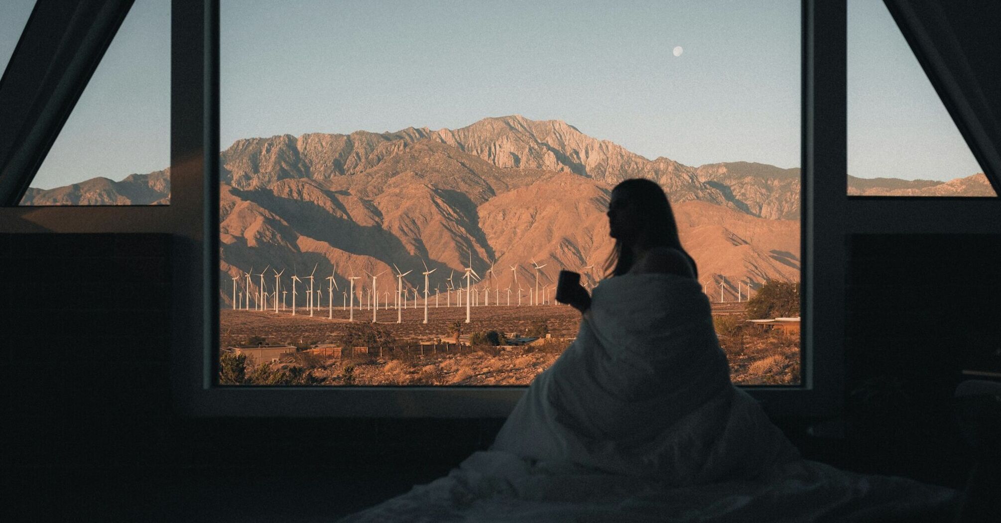 Person wrapped in a blanket looking out a large window at a mountainous landscape with wind turbines