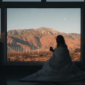 Person wrapped in a blanket looking out a large window at a mountainous landscape with wind turbines