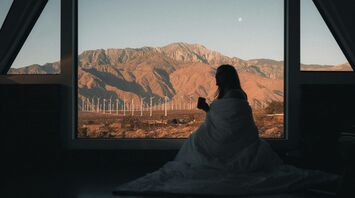 Person wrapped in a blanket looking out a large window at a mountainous landscape with wind turbines