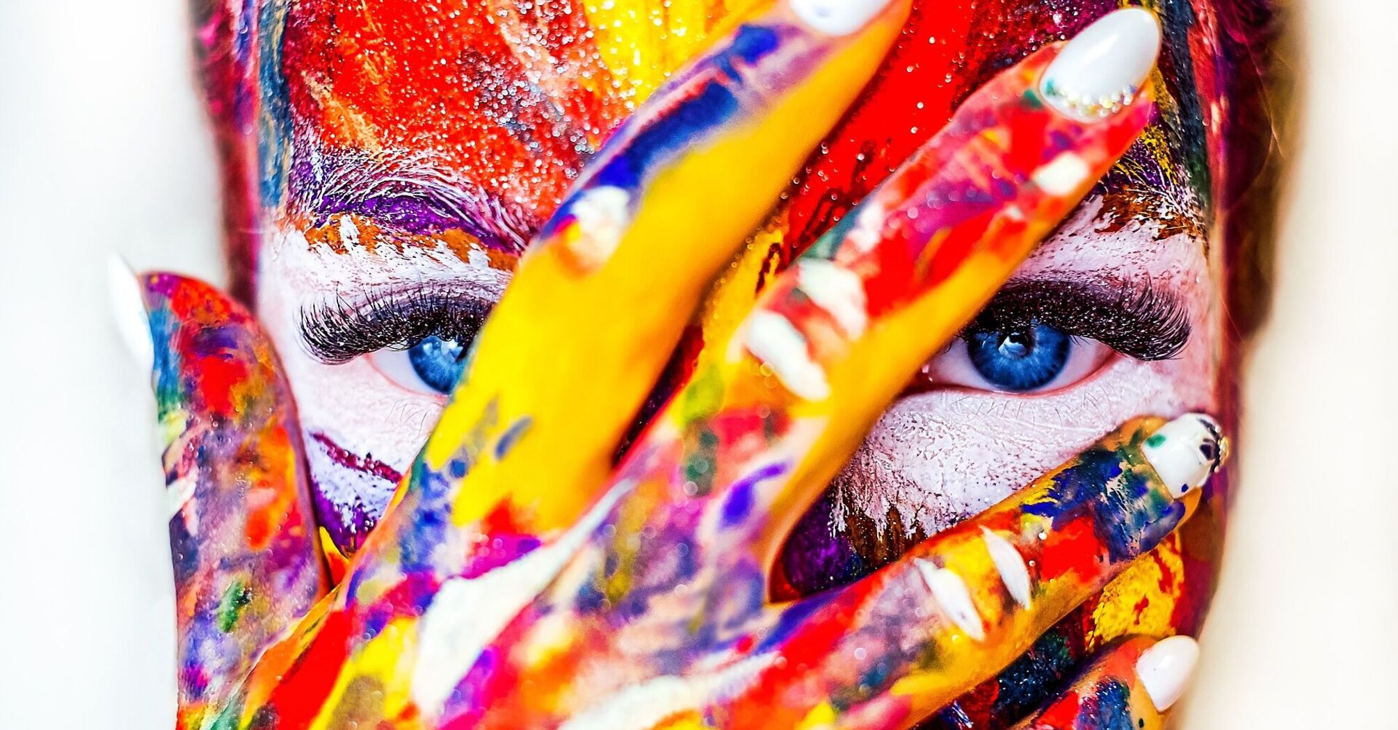 A woman with vibrant paint on her face and hand, peering through colored fingers